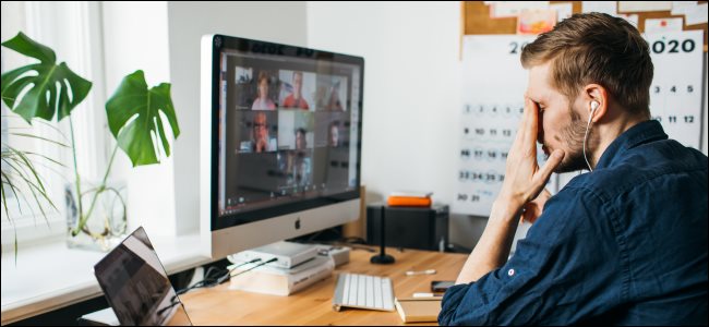 A man with his hand on his face in front of a Mac desktop and laptop during a Zoom meeting.