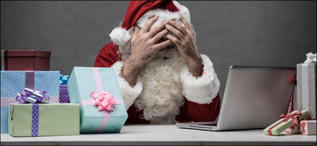 Santa sitting in front of his laptop and a stack of gifts, covering his face with his hands. 