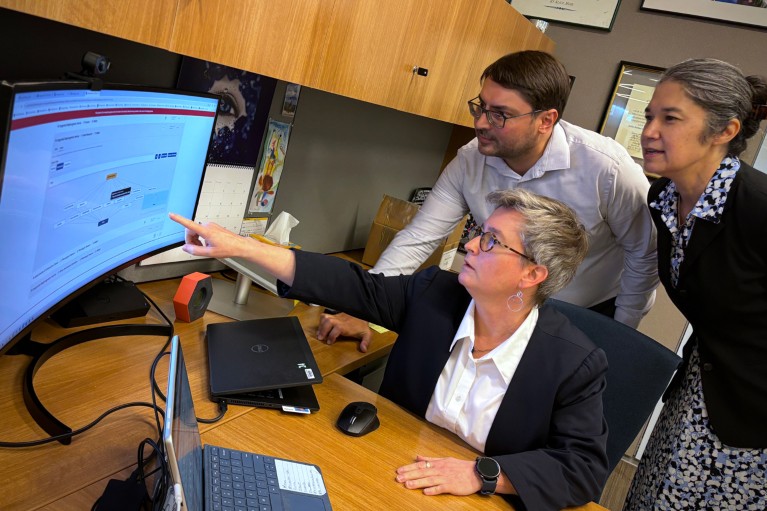 Joni Rutter sits at a desk and points to a computer screen with two colleagues standing behind