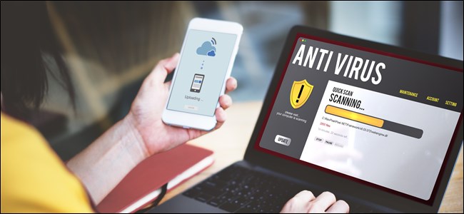 A hand holding a smartphone that's running an antivirus, next to a laptop on a desk that's also running an antivirus scan.