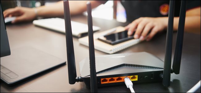 A wireless router on a table with a man using a Mac desktop computer and iPhone in the background.,